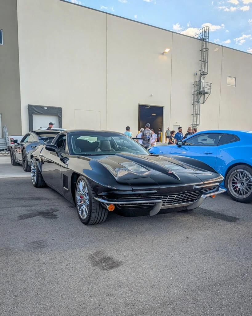 Black Chevrolet Corvette parked beside a blue Ford Mustang and another car in a lot with people and a building in the
