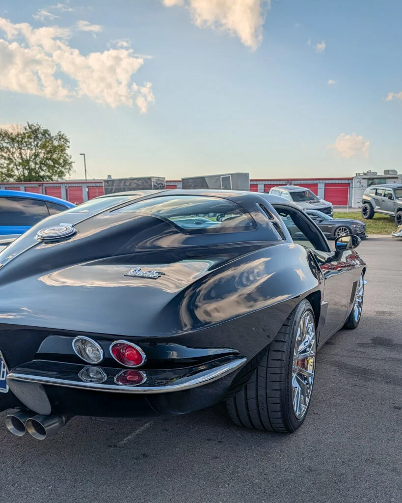 Black Chevrolet Corvette parked outdoors with trailers and other vehicles in the background under a partly cloudy sky.