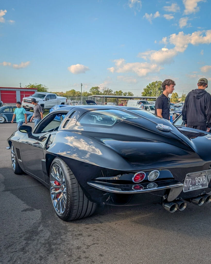 Black Chevrolet Corvette with rear window louvers and quad exhaust tips at an outdoor car event with people nearby
