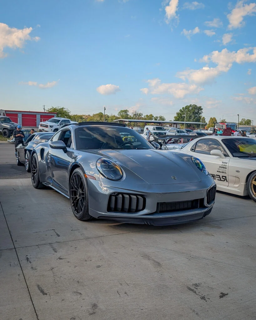 Silver Porsche 911 parked at a car event with other vehicles and people in the background under a blue sky.