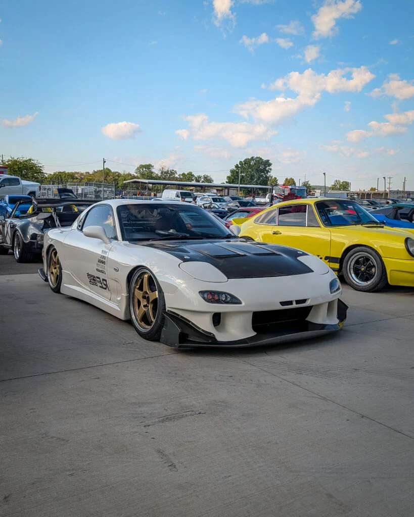 White Mazda RX-7 with black hood parked next to a yellow Porsche 911 in an outdoor car event lot under clear sky
