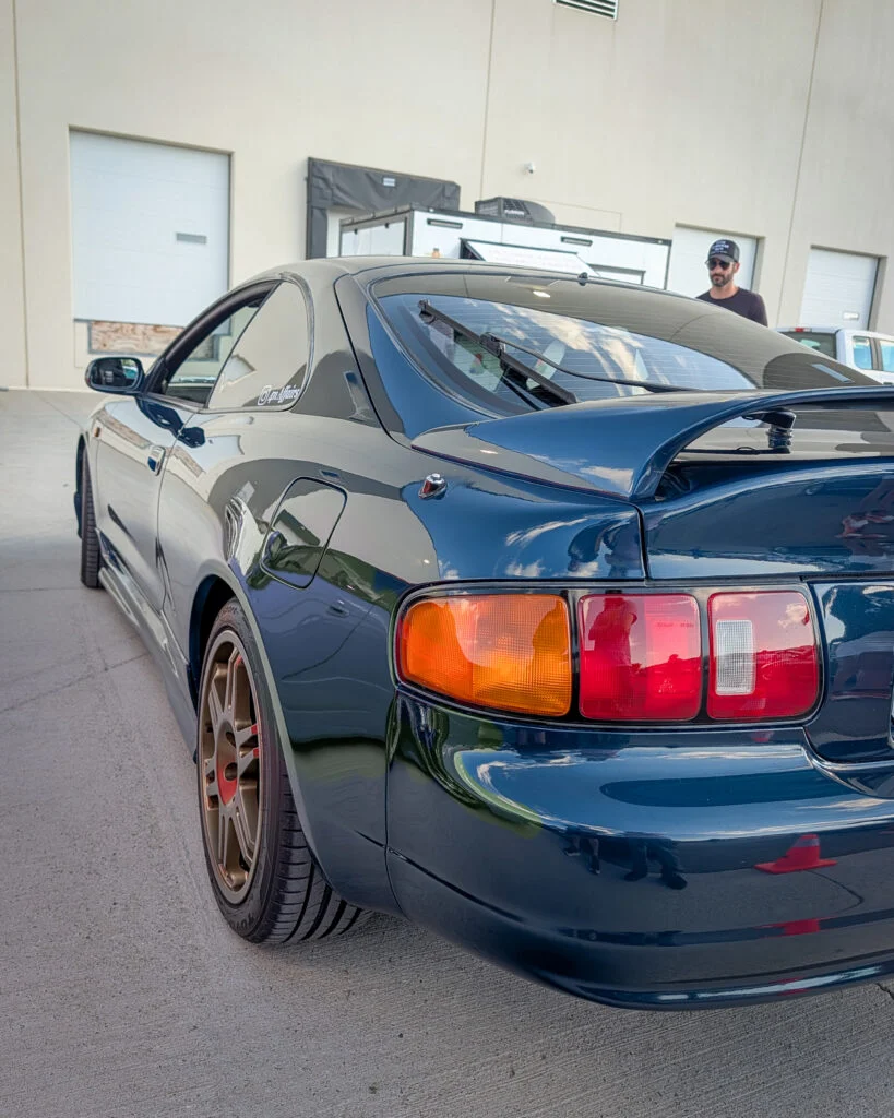 Dark blue Toyota Celica GT-Four with rear spoiler and bronze wheels parked outside a beige industrial building