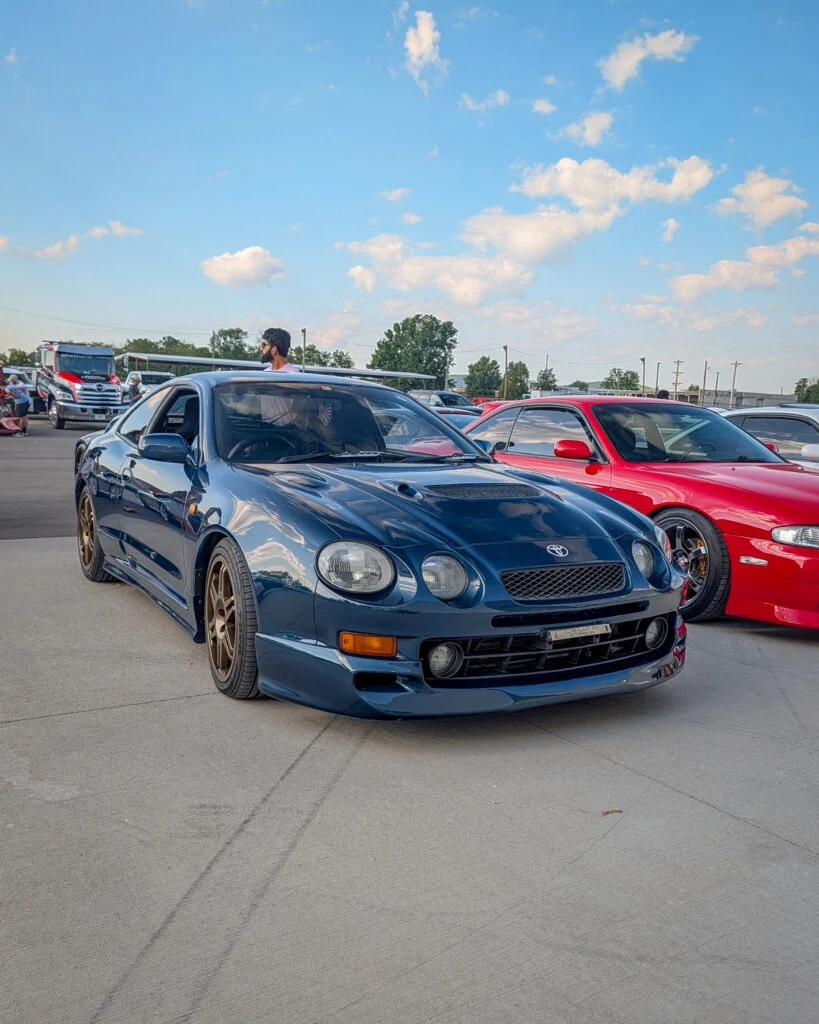 Dark blue Toyota Celica GT-Four parked outdoors with a red coupe beside it and people in the background under a partly