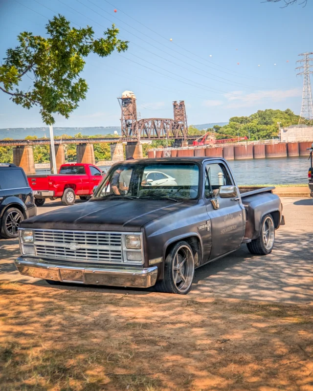 Black Chevrolet Custom Deluxe pickup truck parked near a river with a rusty metal bridge and power lines in the background
