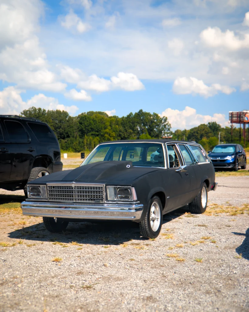 Black Chevrolet Malibu wagon with raised hood parked outside on gravel with blue sky and some other vehicles in background