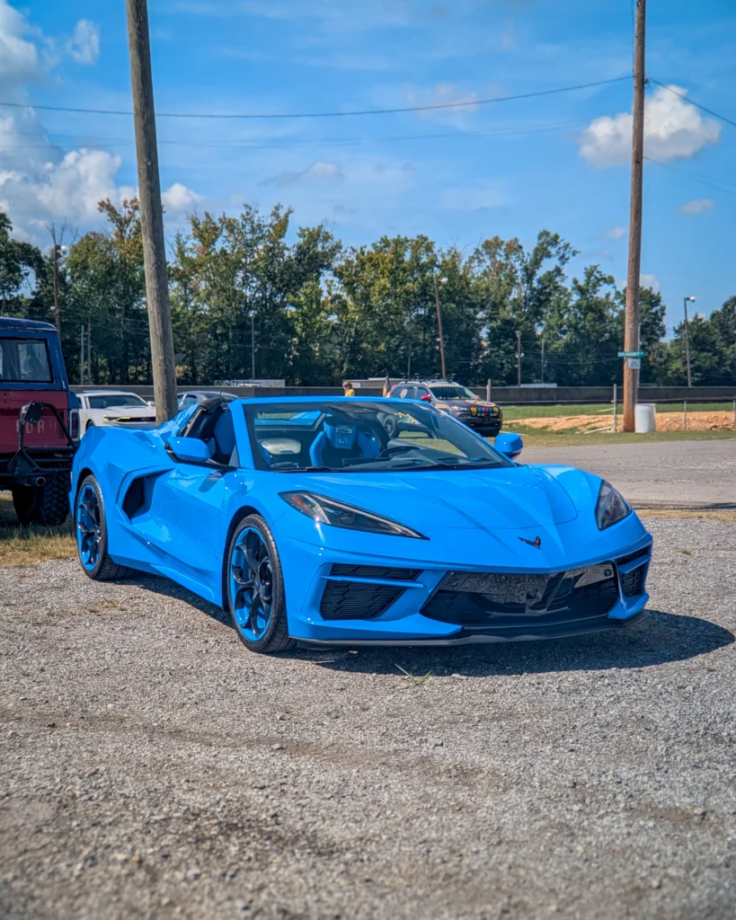 Blue Chevrolet Corvette convertible parked on gravel with trees and a utility pole in the background.