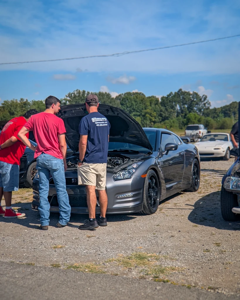 Gray Nissan GT-R with open hood and three men standing and looking at the engine in an outdoor car meet setting