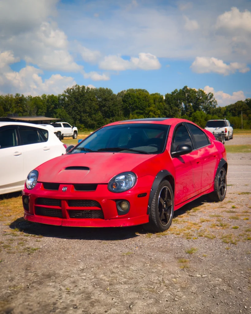 Red Dodge Neon coupe parked on gravel lot with white vehicles and green trees in background