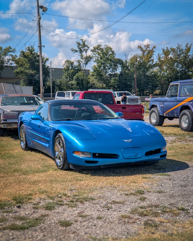 Blue Chevrolet Corvette parked on grass with other vehicles and people in background at a car event