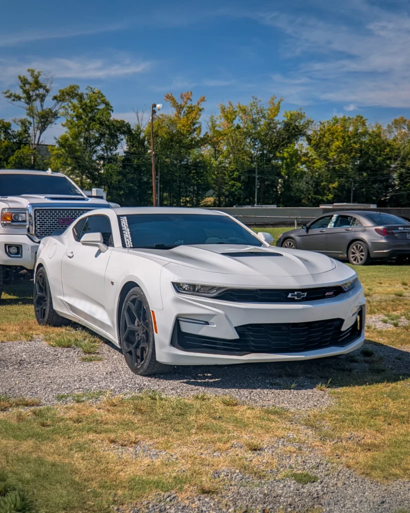 White Chevrolet Camaro SS parked on gravel and grass with trees and other vehicles in the background under a blue sky