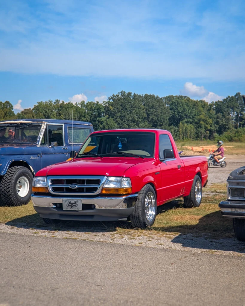 Red Ford Ranger pickup parked outdoors beside older blue Ford Bronco with trees and motorcyclist in background