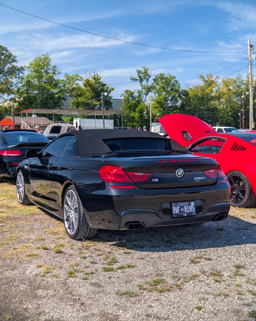 Black BMW 650i convertible parked at an outdoor car event with a red car and other vehicles in the background.