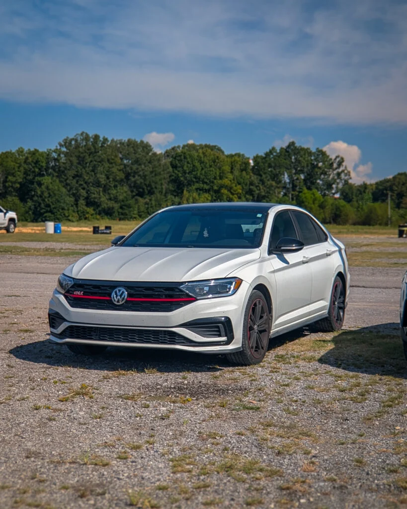 White Volkswagen Jetta GLI parked on gravel with trees and blue sky in background.