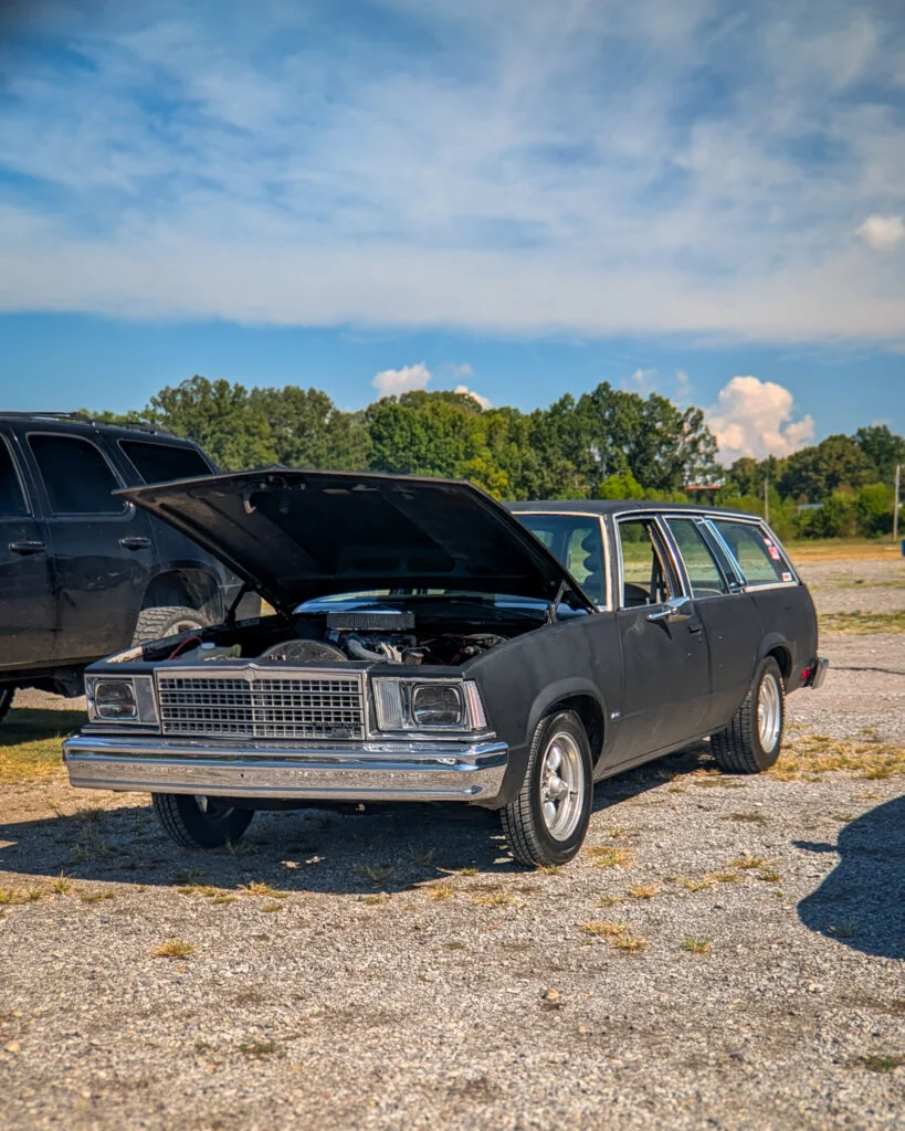 Black Chevrolet Malibu wagon with hood open at an outdoor car event under a blue sky