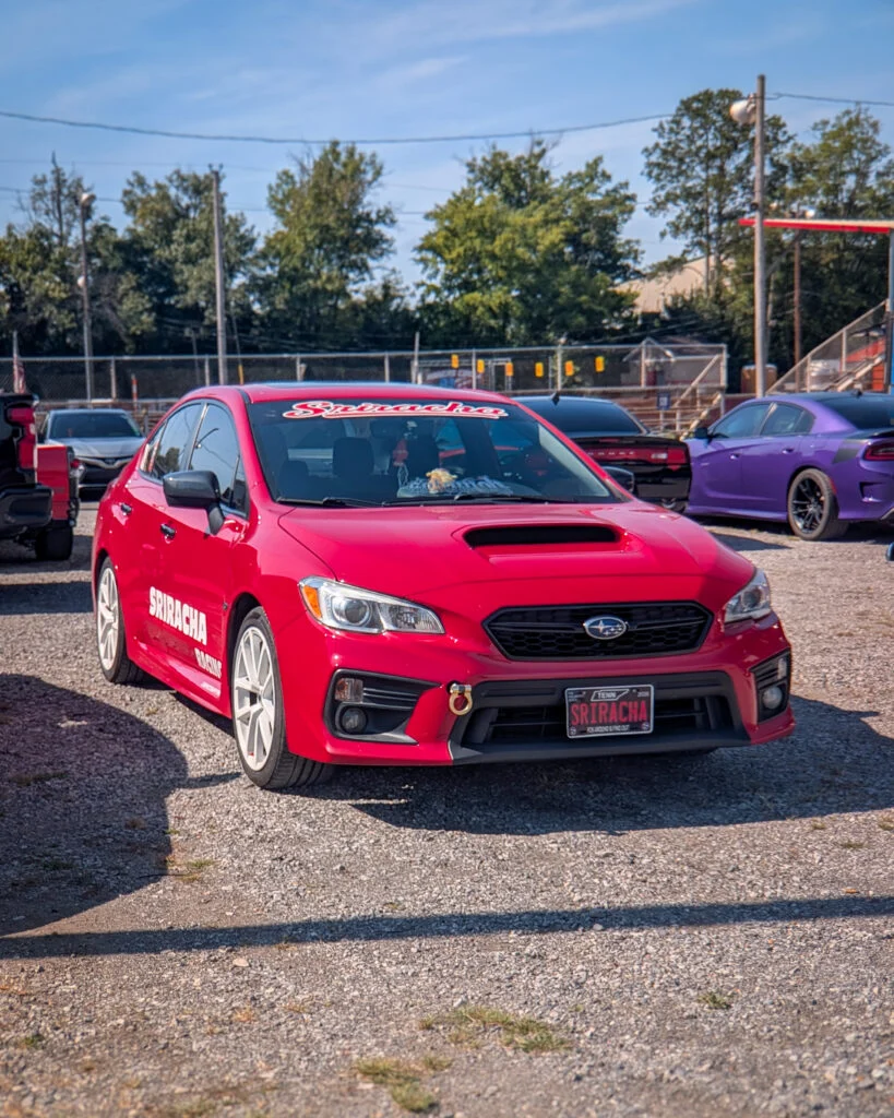 Red Subaru WRX sedan with Sriracha Racing decals parked outdoors with other vehicles and trees in the background