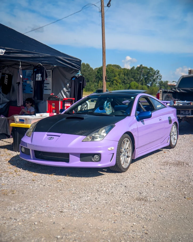Lilac Toyota coupe with black hood parked near a pop-up tent at an outdoor event on a sunny day