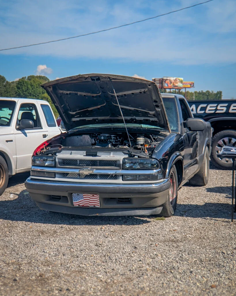 Black Chevrolet S-10 with hood open parked outdoors on gravel next to a white SUV under clear sky