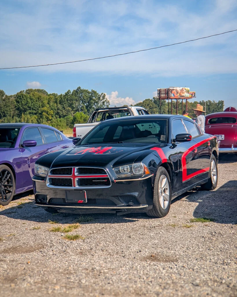 Black Dodge Charger with red decals parked on gravel with other vehicles and people in the background.