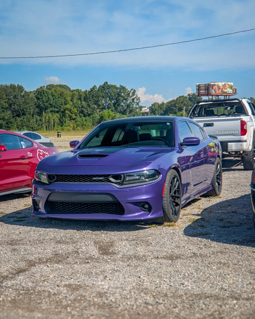 Purple Dodge Charger Daytona parked in a lot with trees and a clear sky in the background, with parts of other vehicles