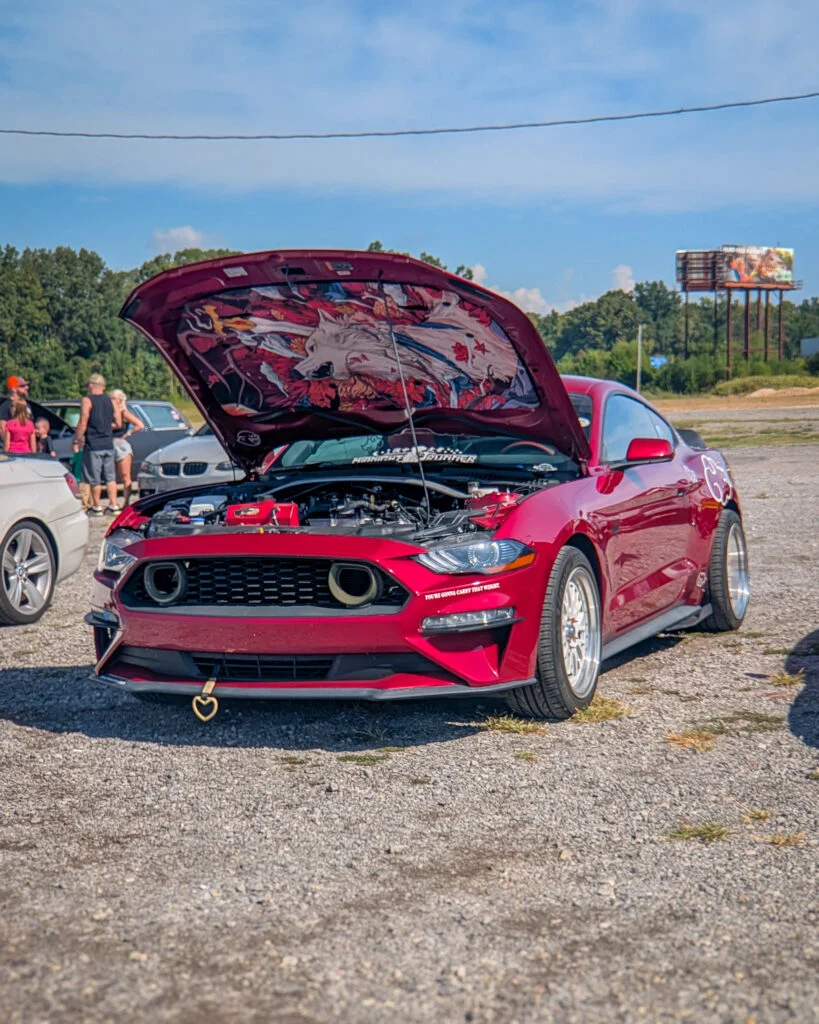 Red Ford Mustang with hood open showing artwork underneath at an outdoor car event during daytime