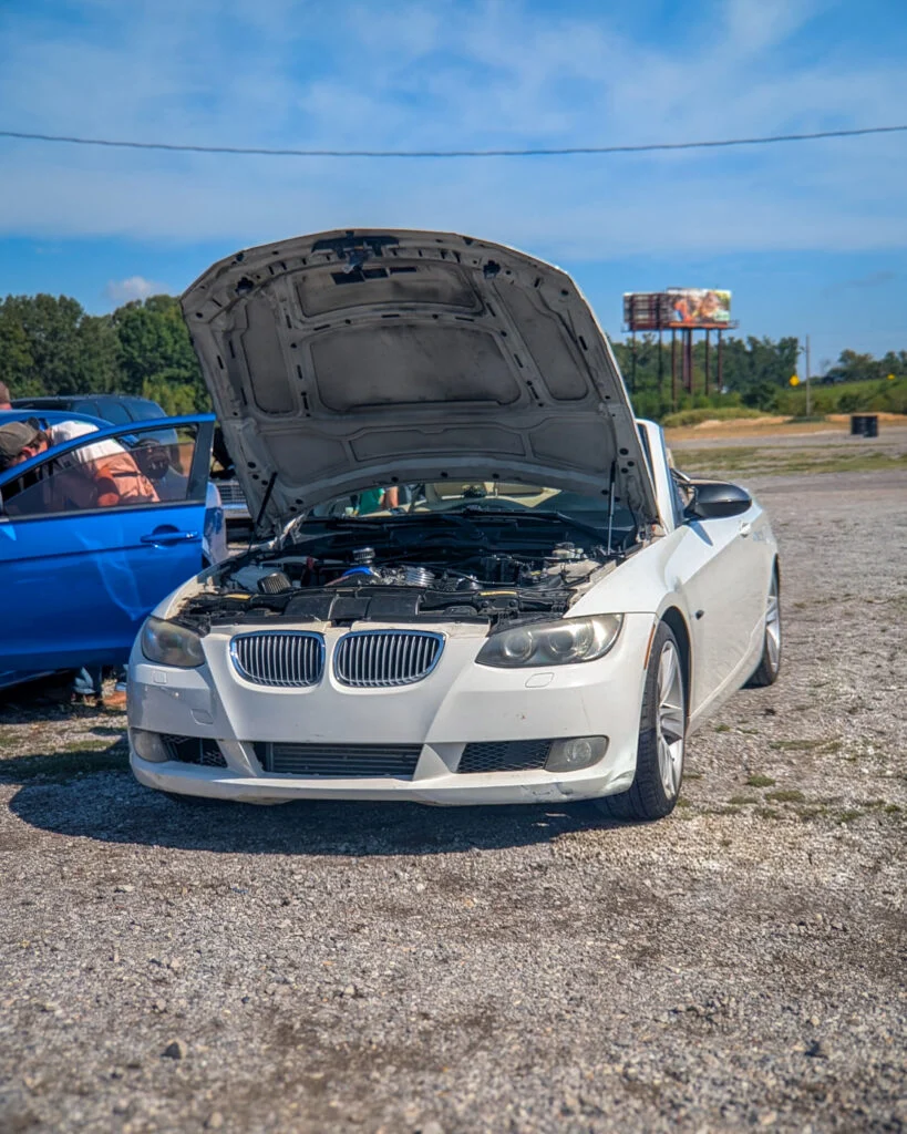 White BMW sedan with hood open in outdoor parking lot with blue sky and another vehicle partly visible