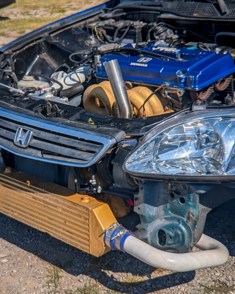 Close-up of a Honda Civic engine bay with a blue VTEC valve cover and a gold intercooler at a car event outdoors.
