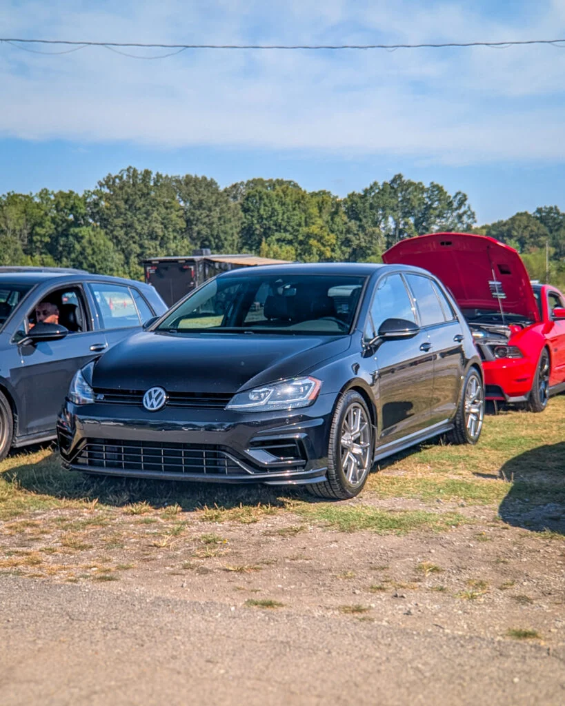 Black Volkswagen Golf R parked on grass with red car with open hood behind it at outdoor car event