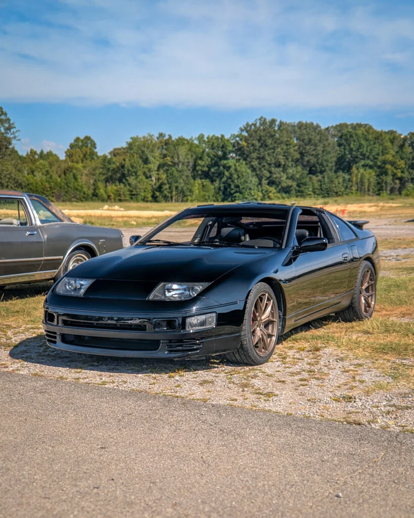 Black Nissan 300ZX and partial view of gray coupe parked outdoors on a clear day with trees in background