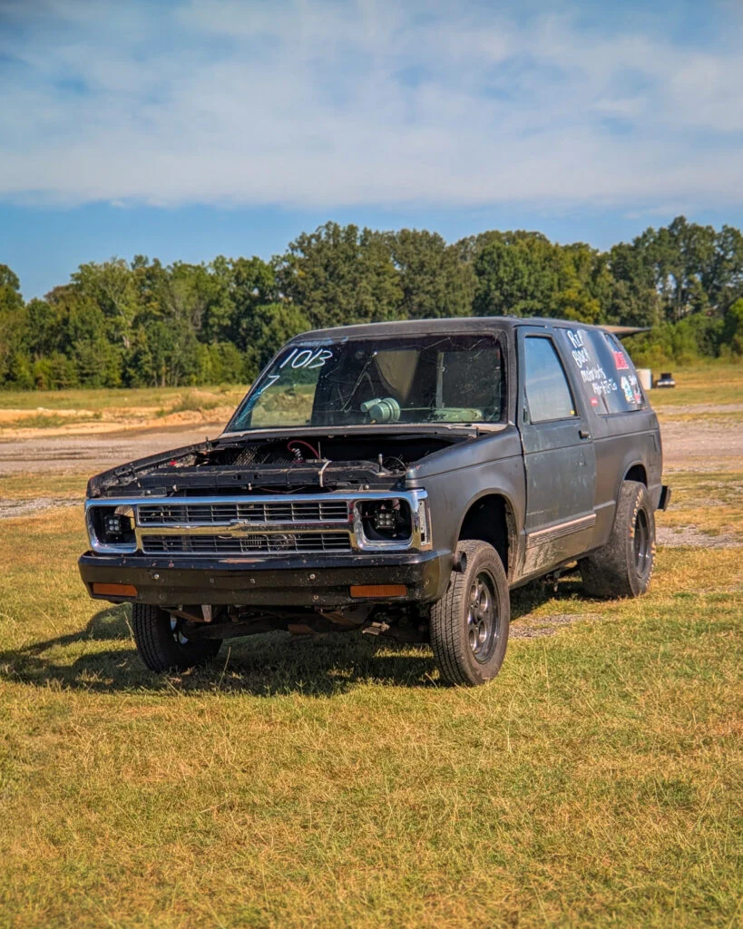 Black Chevrolet S-10 Blazer without hood and broken windshield parked on grass under blue sky with trees in background