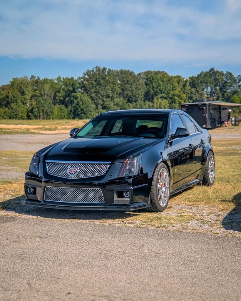 Black Cadillac CTS-V sedan parked on gravel area with trees and trailer in background under blue sky