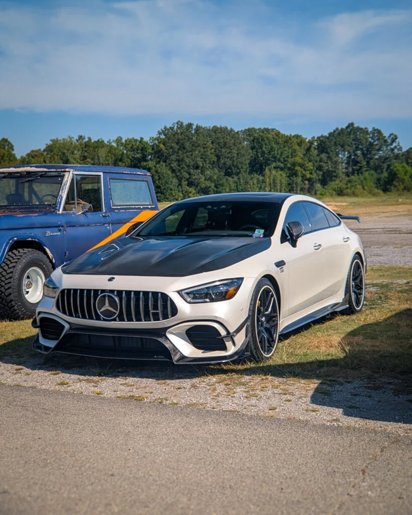 White Mercedes-AMG GT 4-Door Coupe with black hood parked on pavement near an older blue Ford Bronco in a rural area