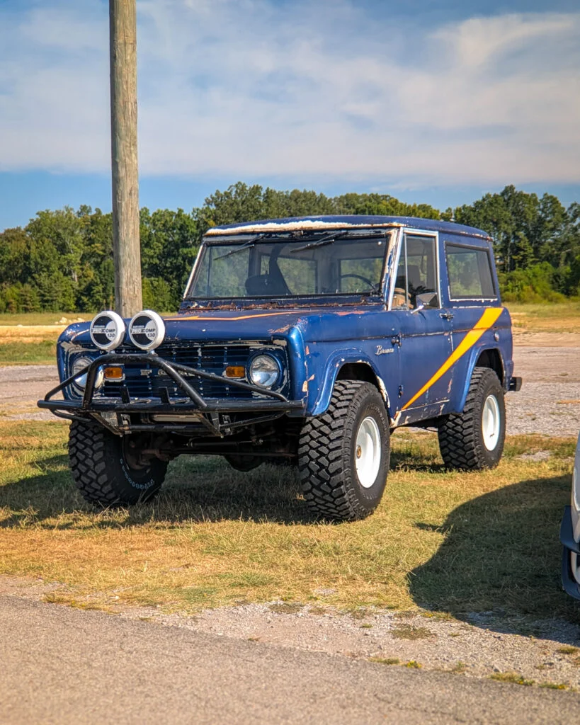 Blue Ford Bronco with orange stripe parked outdoors near a utility pole and trees under a partly cloudy sky