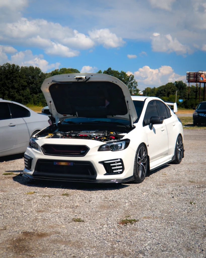 White Subaru WRX STI with hood open, showing engine bay, parked on gravel under a partly cloudy sky with trees in the