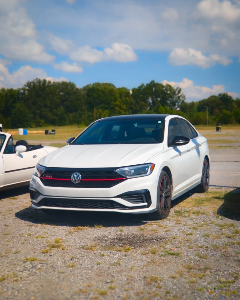 White Volkswagen GLI sedan parked on gravel with trees and blue sky in background