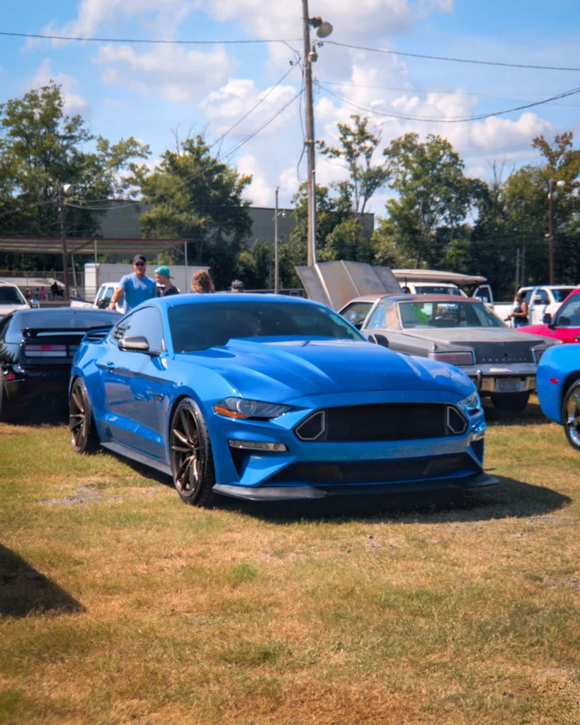 Blue Ford Mustang GT parked on grass at outdoor car event with other vehicles and people in the background
