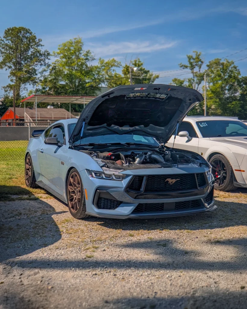 Light blue Ford Mustang GT with hood open and white Dodge Challenger beside it in outdoor parking area