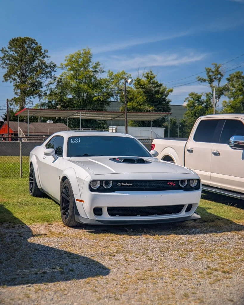 White Dodge Challenger R/T parked on grass and gravel with a silver pickup truck beside it and trees in background