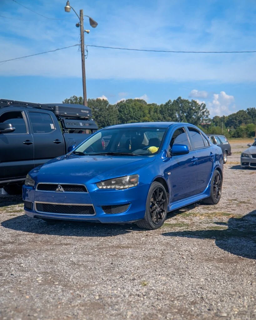Blue Mitsubishi Lancer sedan parked on gravel with other vehicles and trees in background under blue sky.