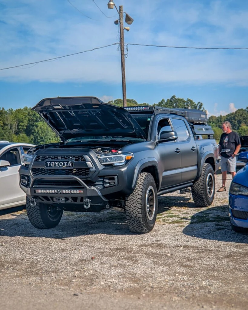 Gray Toyota Tacoma pickup truck with raised hood at outdoor car event with people and other vehicles visible