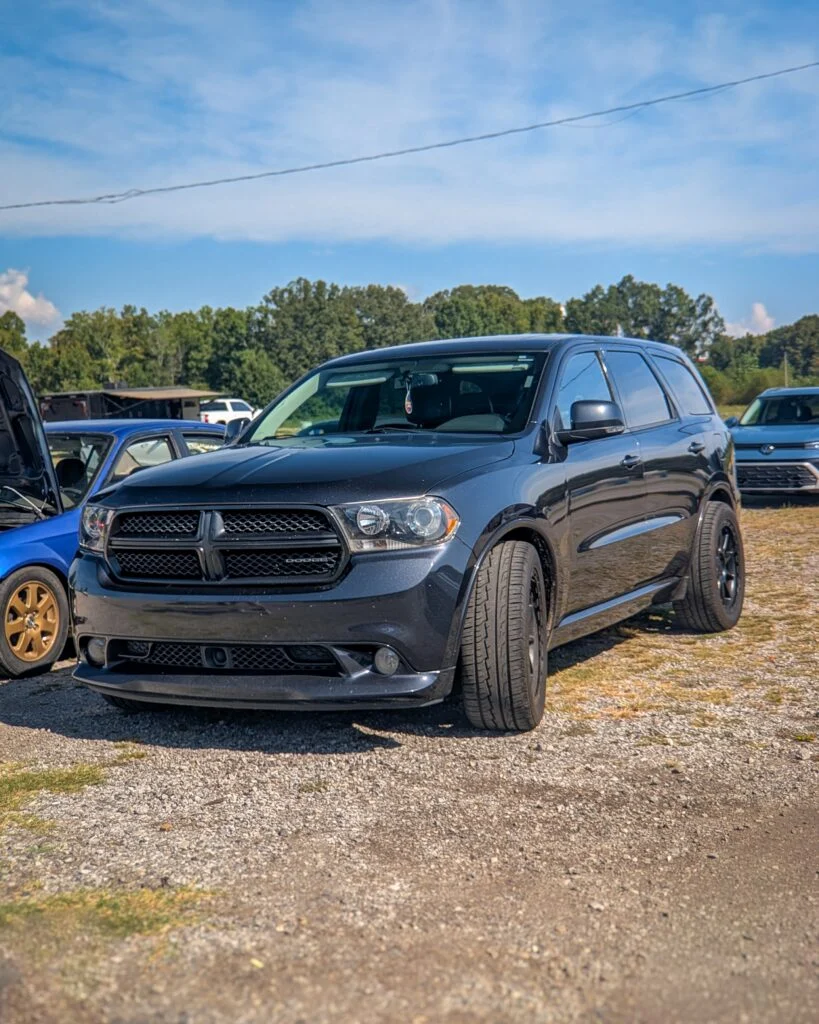 Black Dodge Durango parked on gravel with other vehicles and trees in background under blue sky