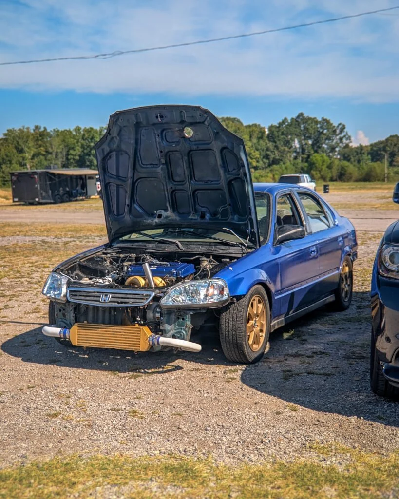 Blue Honda Civic with hood open showing engine bay, parked on gravel with trees and trailer in background