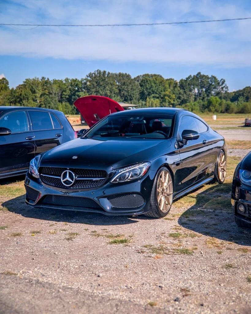 Black Mercedes-Benz AMG C 63 parked outdoors on gravel with a red-hooded vehicle nearby and trees in background