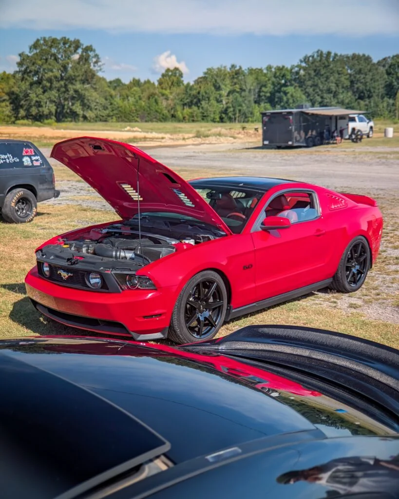 Red Ford Mustang GT coupe with hood open at outdoor automotive event with black vehicle roof reflection in foreground