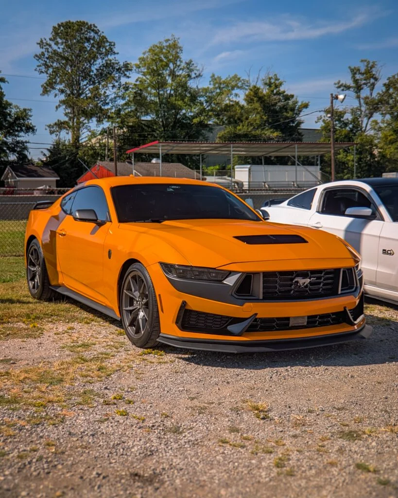 Orange Ford Mustang and white Ford Mustang parked outside on gravel near trees and buildings under clear sky