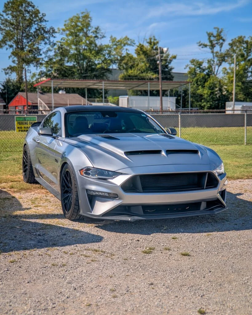 Silver Ford Mustang GT parked on gravel with trees and metal canopy in background