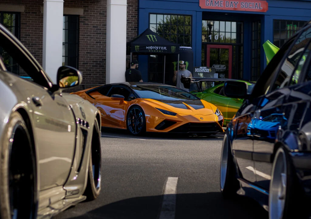 Orange Lamborghini Huracan with black roof parked among other cars in front of a brick building with Monster Energy tent