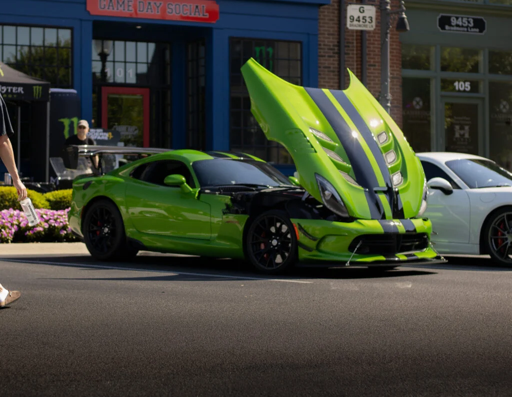 Green Dodge Viper with hood open showing engine at outdoor car event with other cars and buildings in background