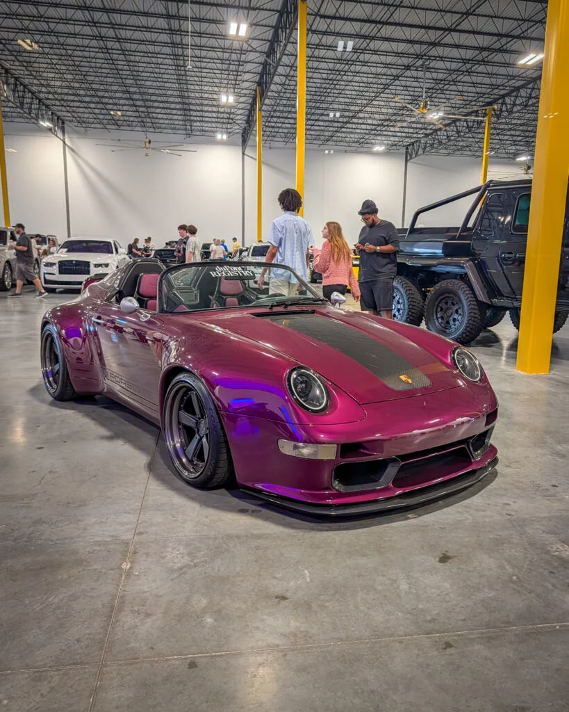 Purple Porsche 911 Speedster with black stripe parked indoors at a car event with several people and other vehicles in the