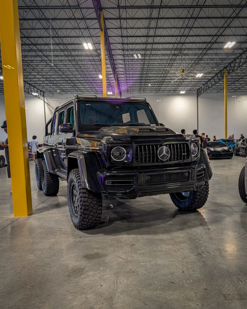 Black Mercedes-Benz G-Class SUV 6x6 in indoor event space with people and other cars in background