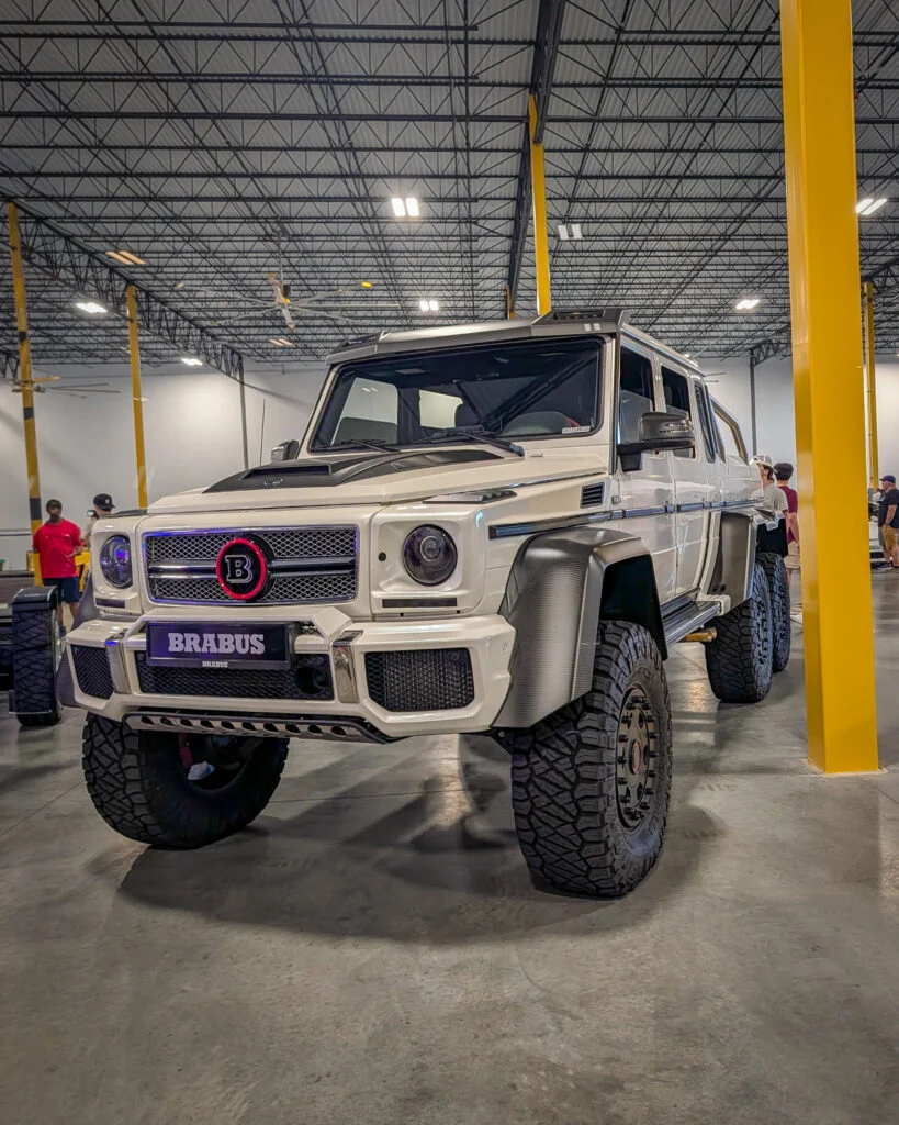 White Brabus Benz G-Class 6x6 with off-road tires in an indoor event space with yellow columns and people in the background.
